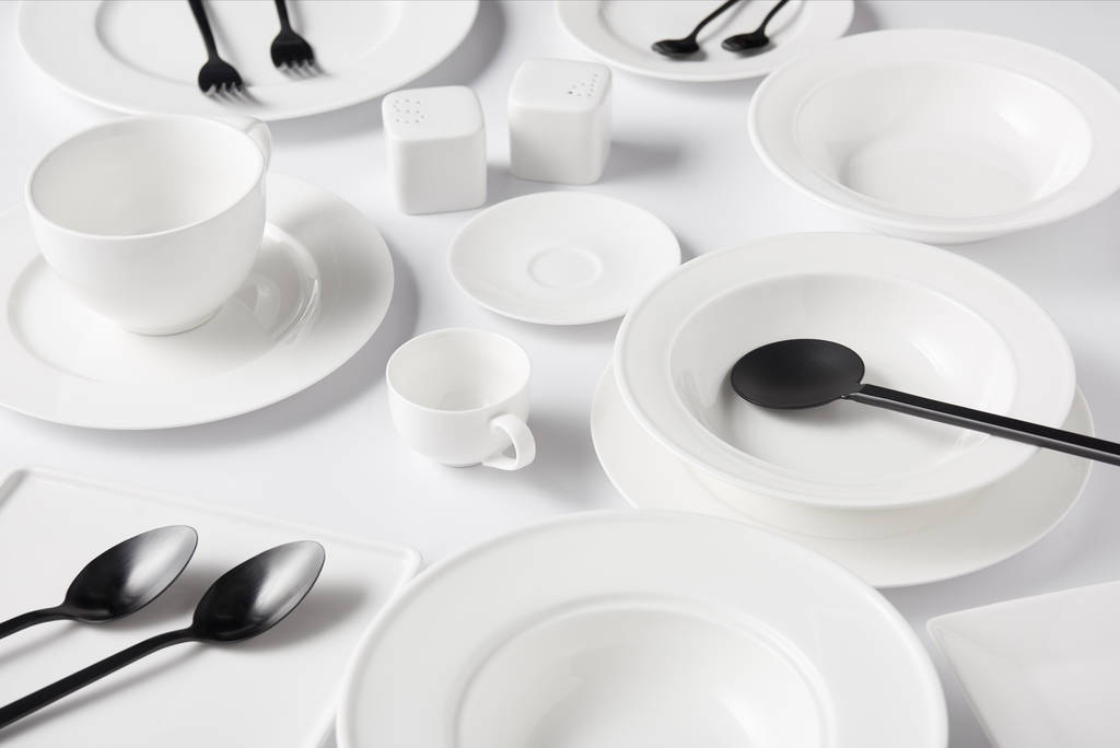 enfoque selectivo de diferentes platos, taza, tazón, cuchara de ensalada, bodega de sal y echador de pimienta, tenedores con cucharas en la mesa blanca
 - Foto, imagen