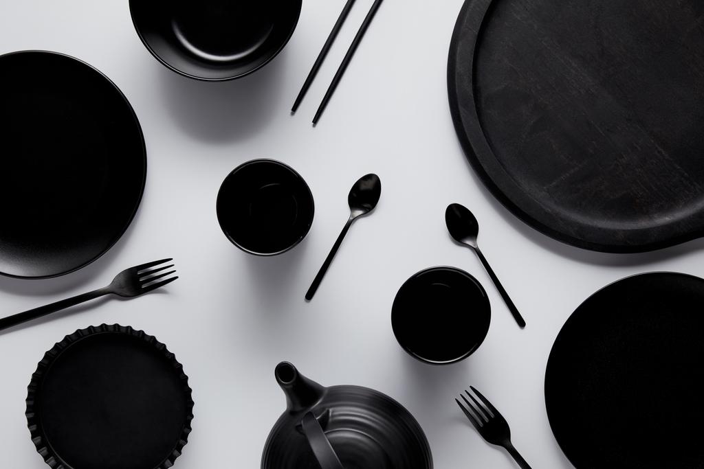vista superior de bule preto, colheres, garfos, pauzinhos, copos, bandeja, tigela, pratos e assadeira na mesa branca
  - Foto, Imagem