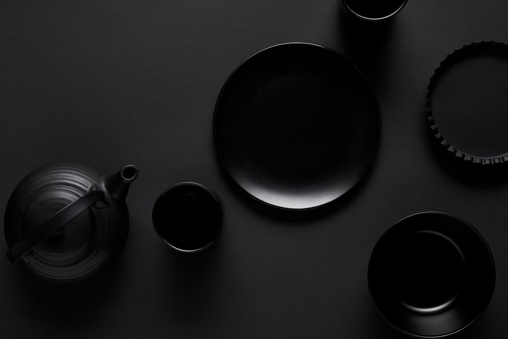 Ansicht von schwarzer Teekanne, Teller, Schüssel, Tassen und Backform auf schwarzem Tisch  - Foto, Bild