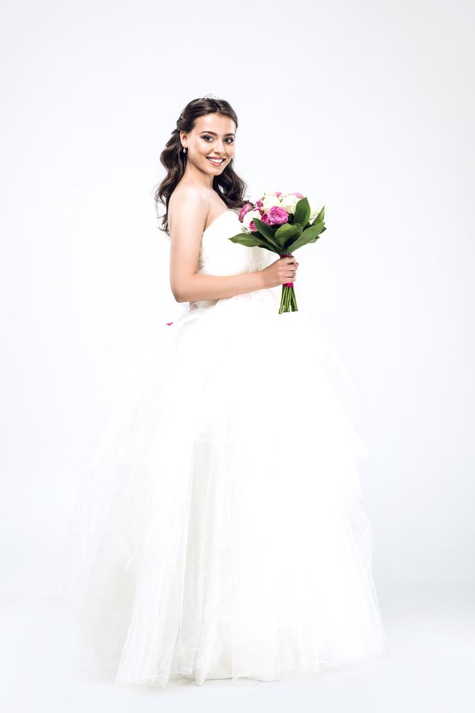 krásná mladá nevěsta ve svatebních šatech s kyticí a při pohledu na fotoaparát na bílém - Fotografie, Obrázek