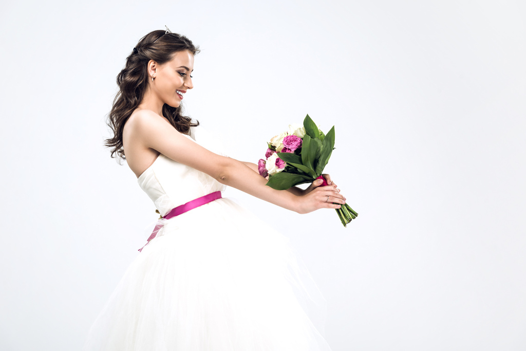 вид сбоку улыбающейся молодой невесты в свадебном платье, держащей свадебный букет изолированный на белом
 - Фото, изображение
