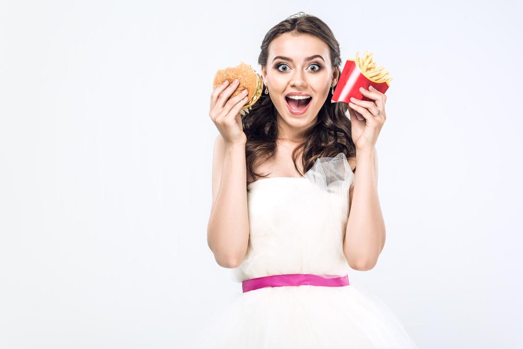 schockierte junge Braut im Hochzeitskleid mit Burger und Pommes frites blickt isoliert in die Kamera auf Weiß - Foto, Bild