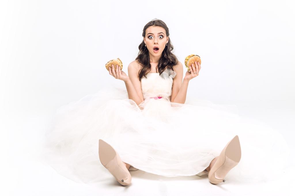shocked young bride in wedding dress sitting on floor with burgers in hands on white - Photo, Image