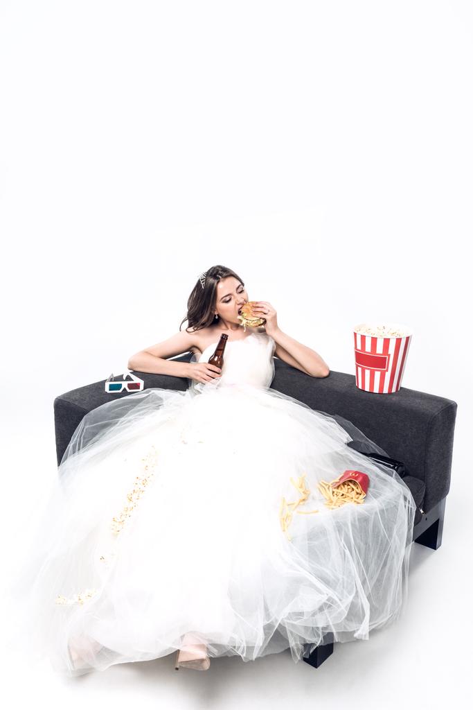 hongerige jonge bruid in trouwjurk zittend op de Bank met bier en junkfood op wit - Foto, afbeelding