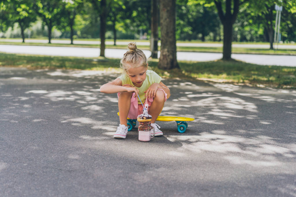 Selektiver Fokus des kleinen Kindes, das auf dem Skateboard sitzt und auf der Straße Dessert isst - Foto, Bild