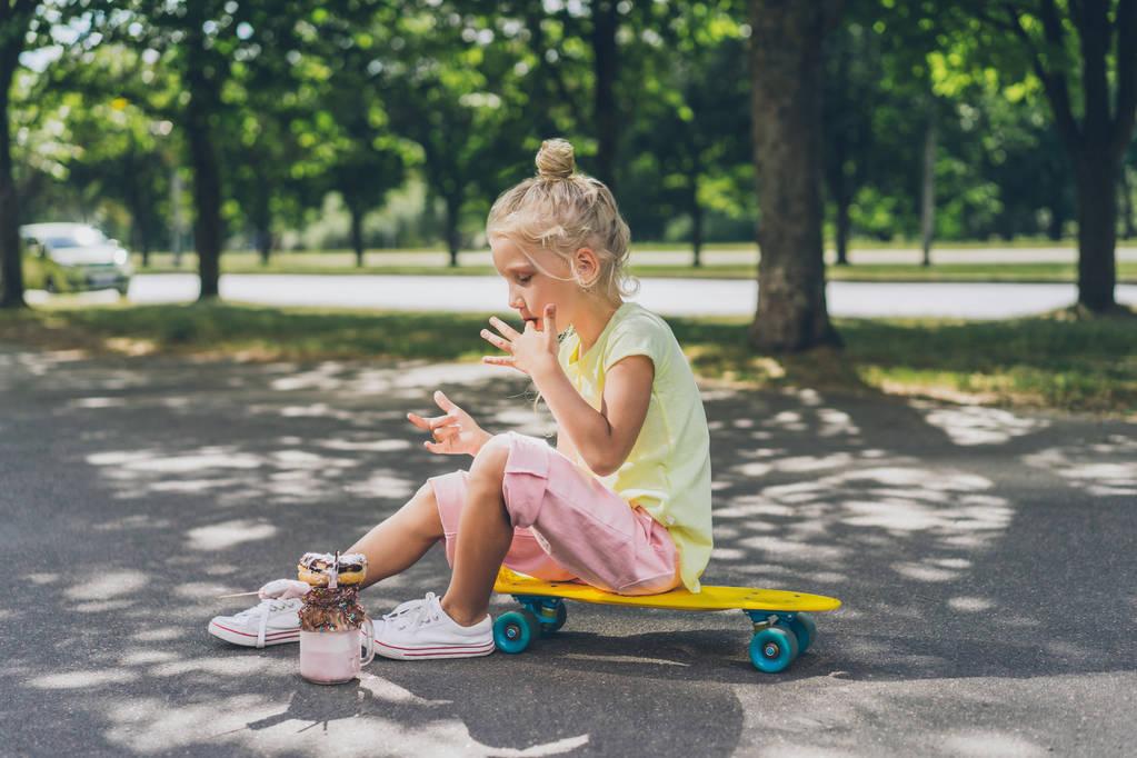 Seitenansicht eines kleinen Kindes, das sich die Finger vom Dessert leckt und auf dem Skateboard in der Stadtstraße sitzt - Foto, Bild