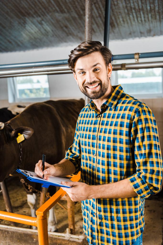 granjero guapo revisando vacas en establo y sujetando portapapeles con documentos
  - Foto, imagen