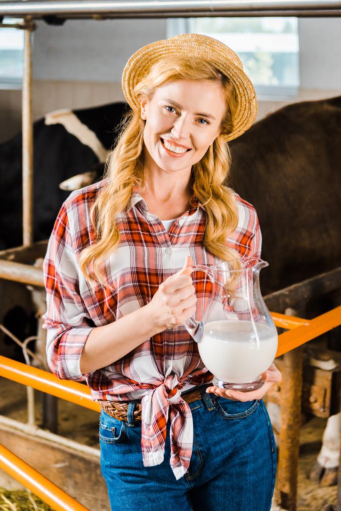 retrato de sonriente agricultor atractivo sosteniendo jarra de leche en establo y mirando a la cámara
 - Foto, Imagen