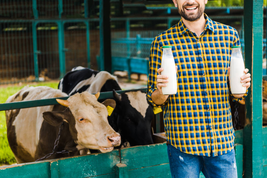 körülvágott kép a mosolygó gazdálkodó üveg tehén tejet közelében stabil - Fotó, kép