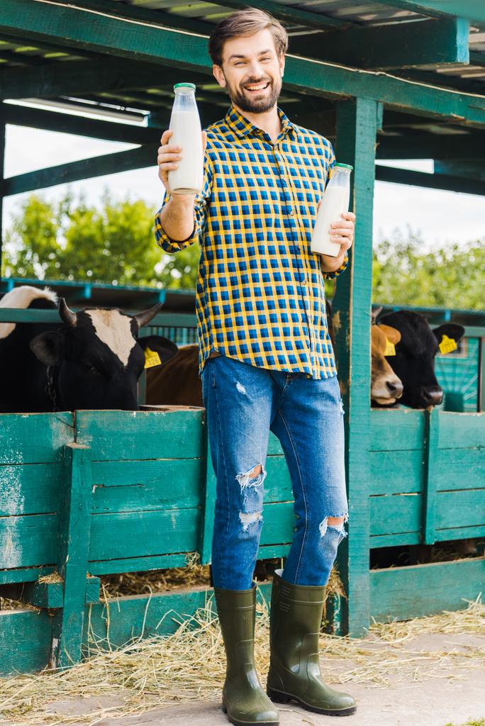 красивий усміхнений фермер тримає пляшки коров'ячого молока біля стабільного
 - Фото, зображення