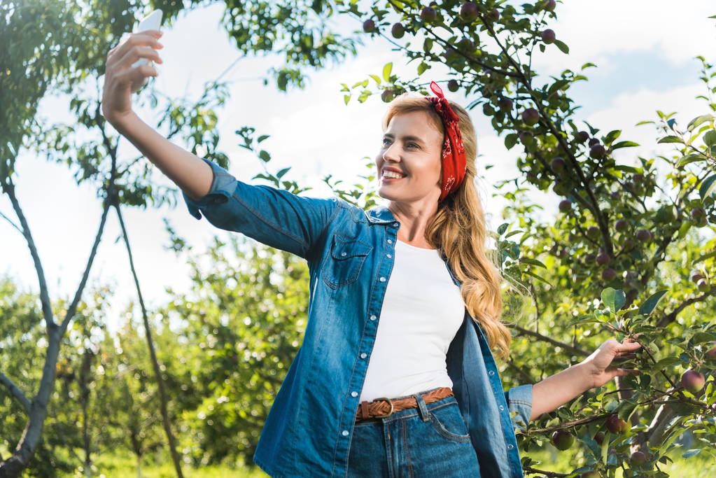 ελκυστική γεωργός λαμβάνει selfie με smartphone στον κήπο της apple στο αγρόκτημα - Φωτογραφία, εικόνα