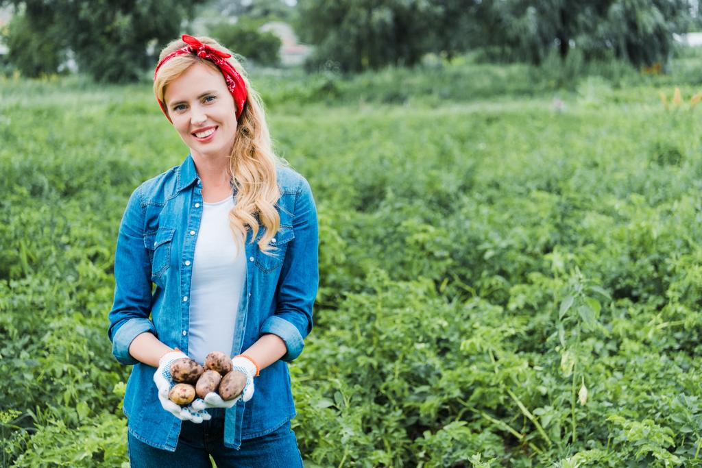 glimlachend aantrekkelijke boer bedrijf rijpe aardappelen in veld op boerderij en camera te kijken - Foto, afbeelding