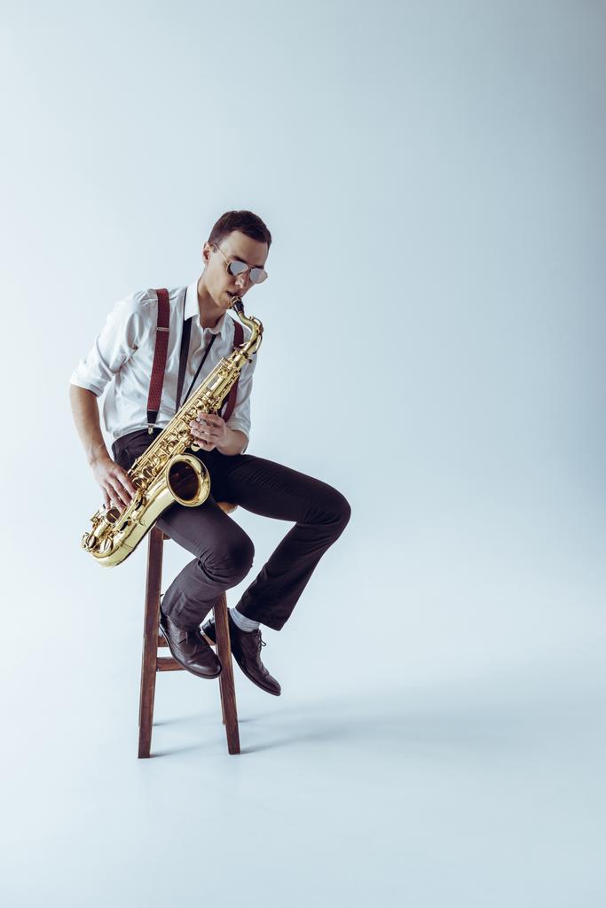 schöner junger Jazzer sitzt auf einem Hocker und spielt Saxofon auf grau  - Foto, Bild