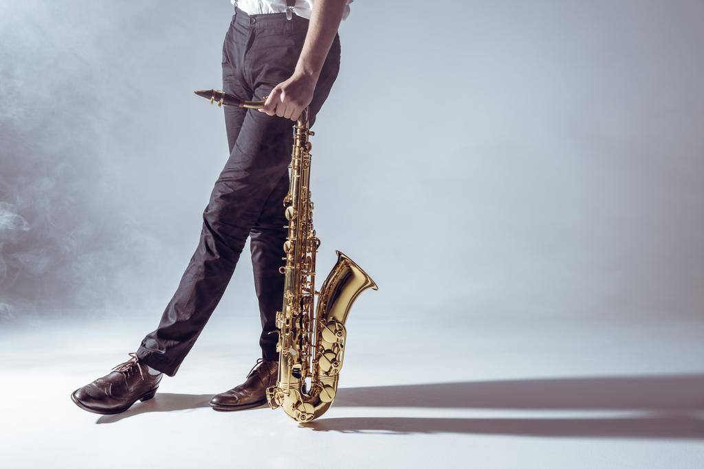 recortado disparo de músico profesional de pie con saxofón en el humo en gris
    - Foto, imagen