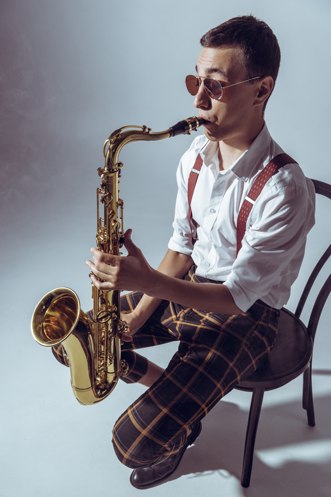 high angle view of stylish young performer sitting on chair and playing saxophone on grey - Photo, Image