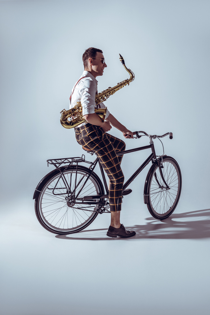 joven músico con estilo en gafas de sol con saxofón y bicicleta en gris
 - Foto, imagen