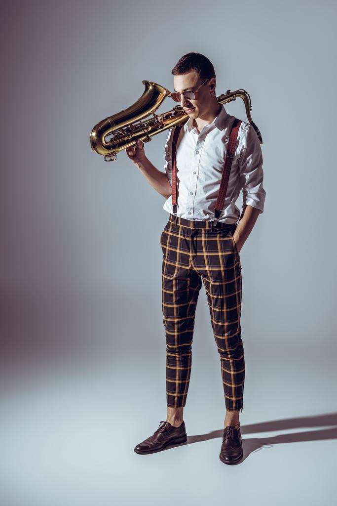 jovem músico elegante em óculos de sol segurando saxofone e olhando para baixo em cinza
 - Foto, Imagem