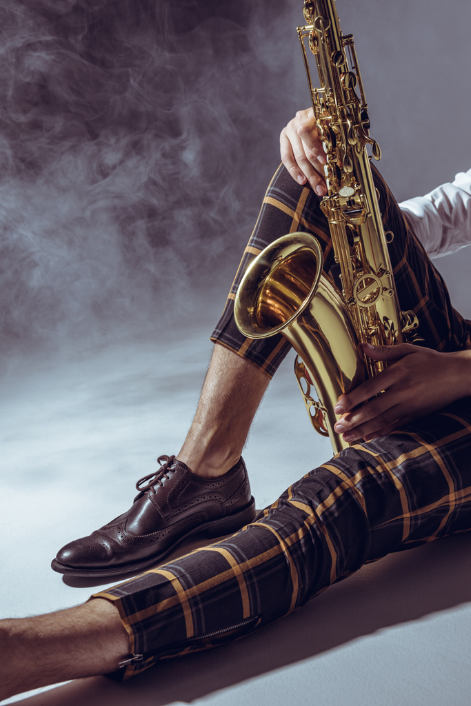 vista parziale di giovane musicista elegante seduto con sassofono in fumo su grigio
  - Foto, immagini