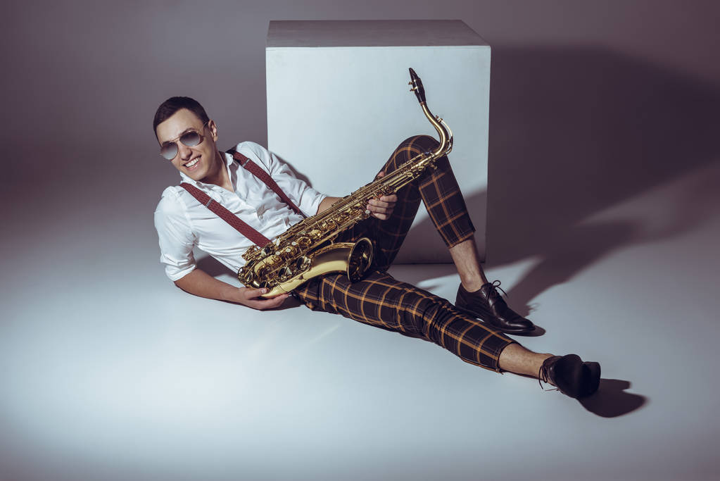 sonriente joven músico en gafas de sol sosteniendo saxofón mientras está acostado en gris
 - Foto, imagen
