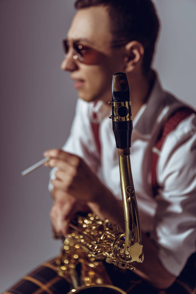 Selektiver Fokus junger Saxofonist beim Zigarettenrauchen auf Grau - Foto, Bild
