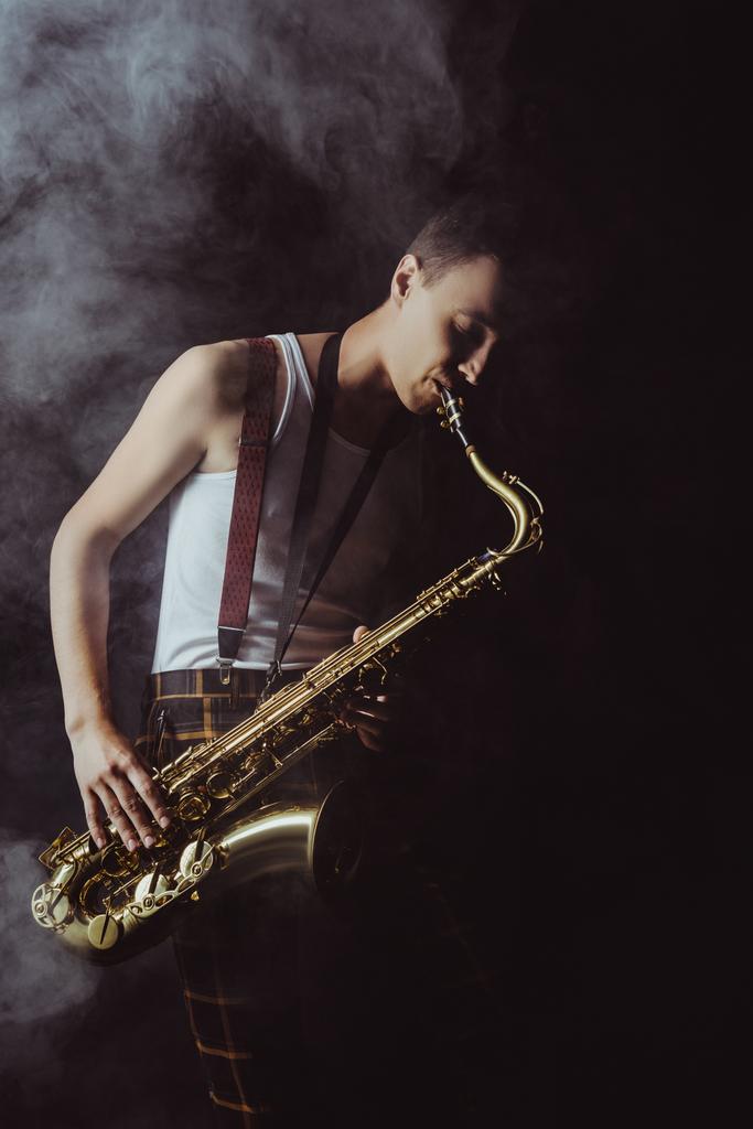 przystojny młody muzyk gra saksofon w dym na czarny - Zdjęcie, obraz