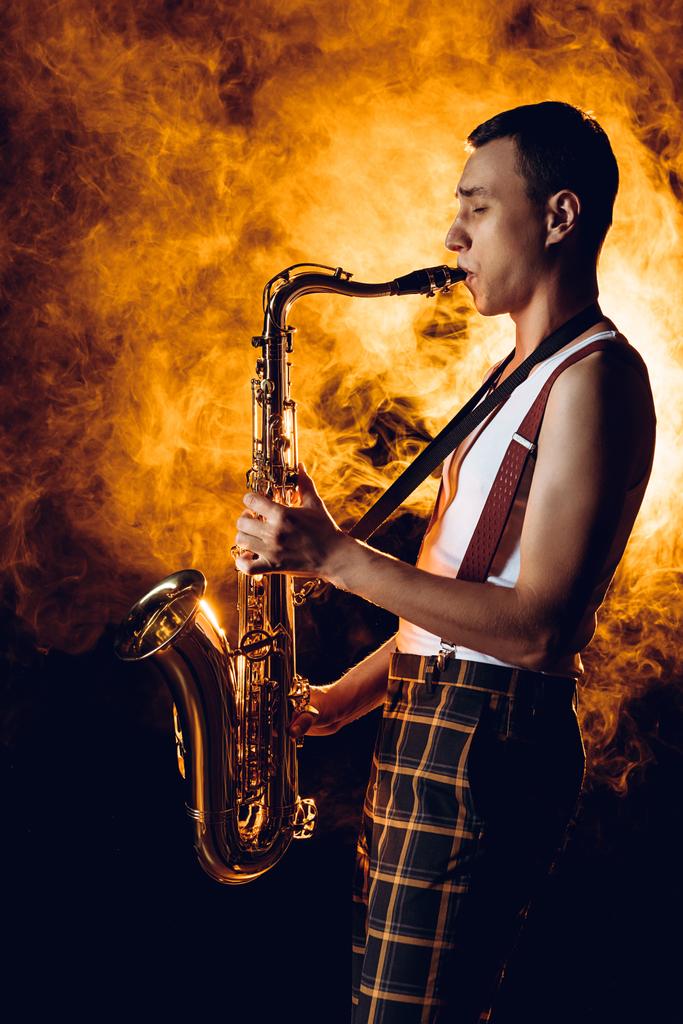 Seitenansicht eines ausdrucksstarken, stilvollen jungen Musikers, der Saxofon in Rauch spielt  - Foto, Bild
