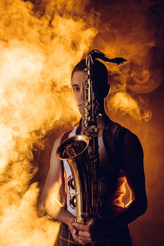 young male musician holding saxophone and looking at camera in smoke - Photo, Image
