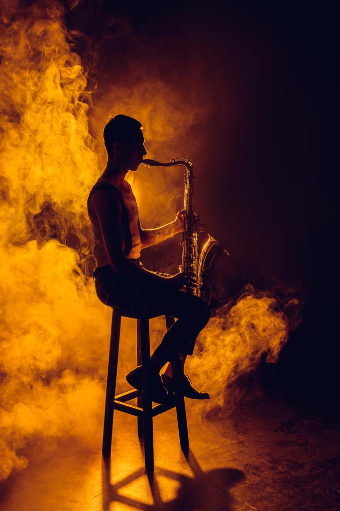 silhueta de jovem músico sentado nas fezes e tocando saxofone na fumaça
  - Foto, Imagem
