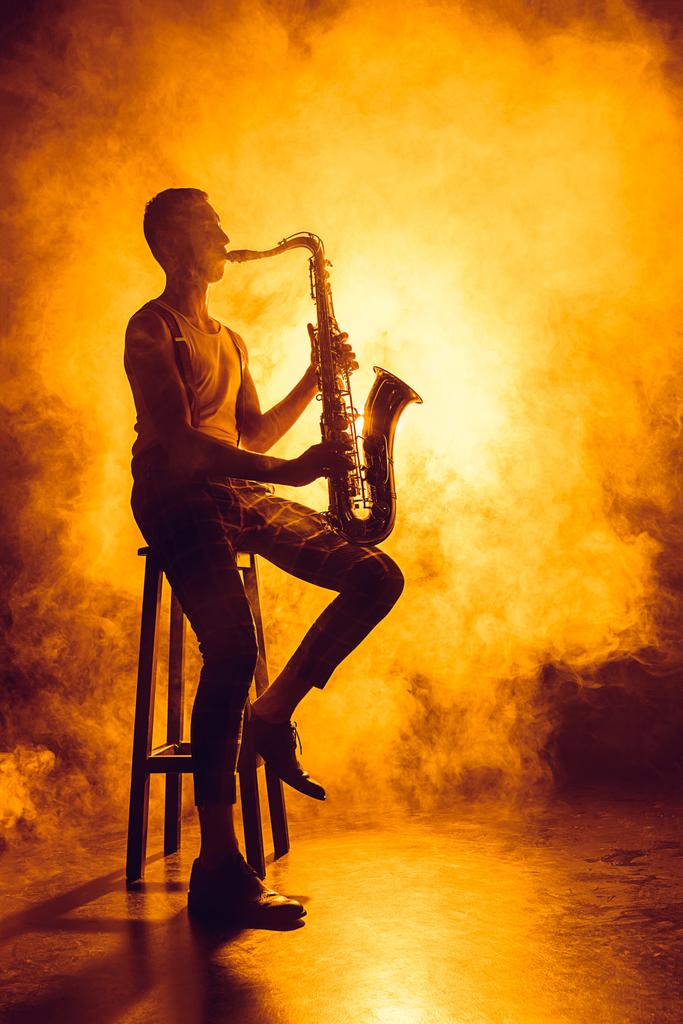 молодий професійний музикант сидить на табуреті і грає в саксофон в диму
  - Фото, зображення