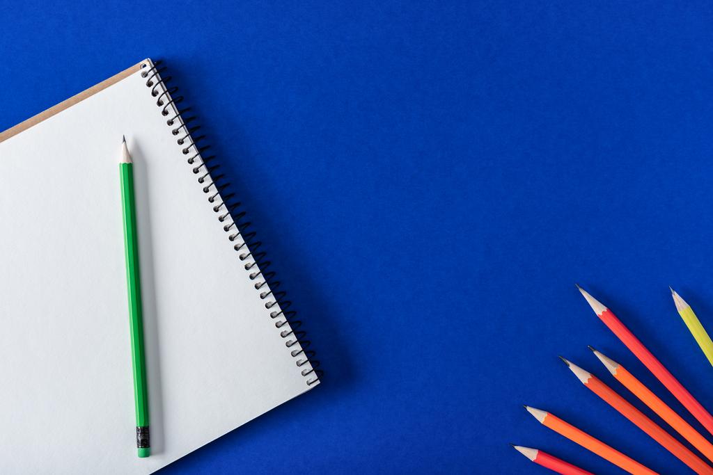 Draufsicht auf arrangierte Buntstifte und leeres Lehrbuch auf blauem Hintergrund  - Foto, Bild