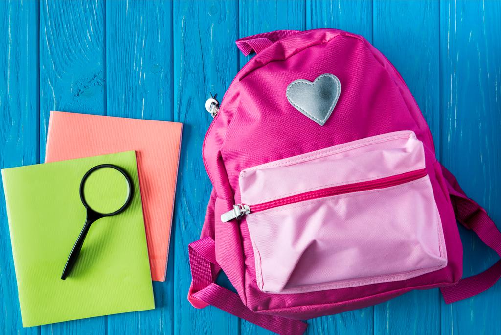 pohled shora učebnic, Lupa a růžový batoh na modré dřevěné pozadí  - Fotografie, Obrázek