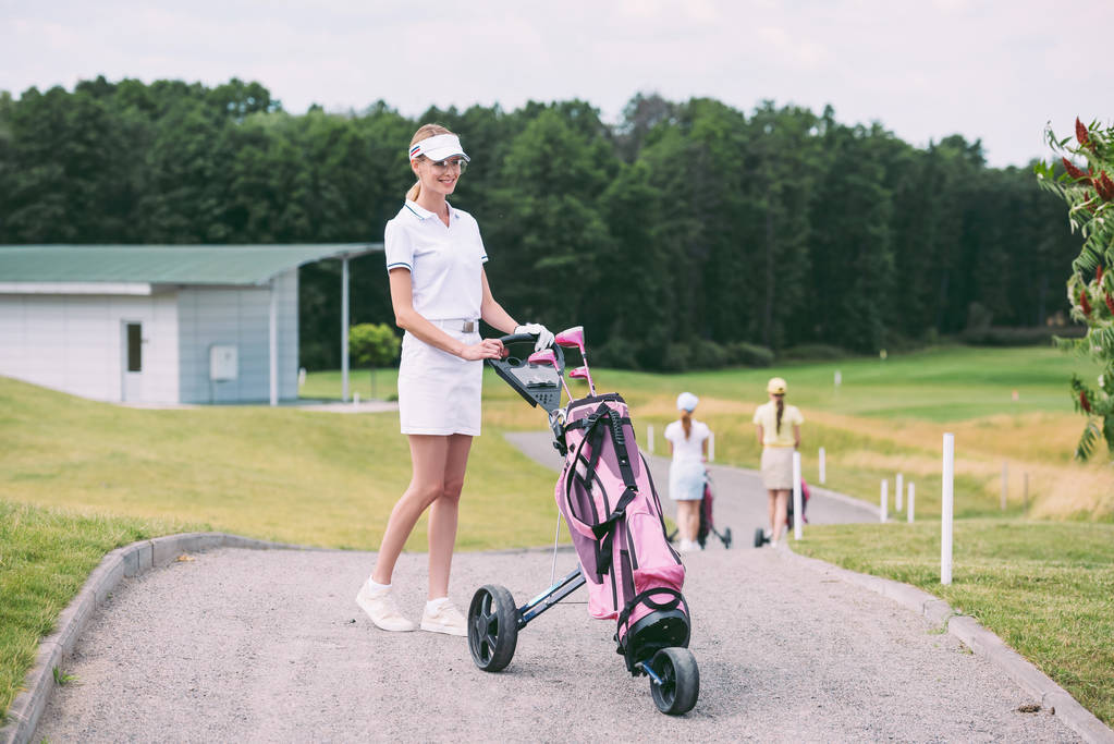 enfoque selectivo de jugadora de golf alegre en gorra y polo blanco con equipo de golf en el campo de golf
 - Foto, imagen
