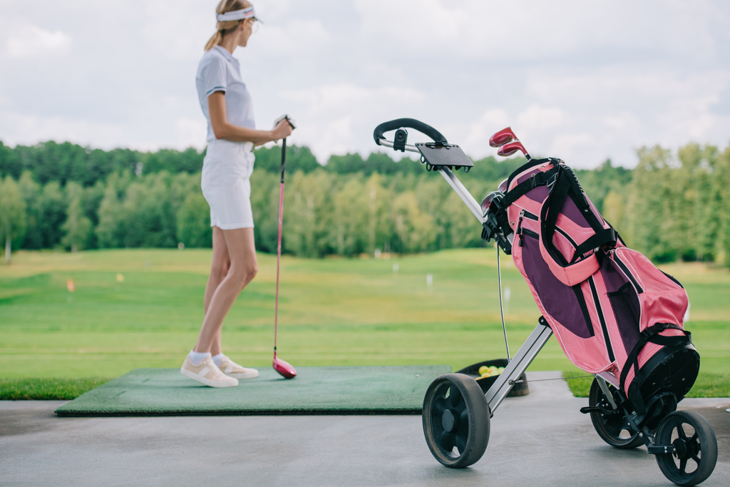 вид сбоку женщины в поло и кепке с клюшкой для гольфа, стоящей на поле для гольфа
 - Фото, изображение