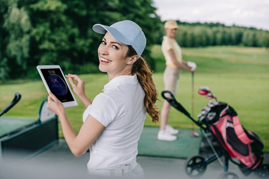 Selektivní fokus usmívající se žena s tabletem a přítel hrát golf za na golfovém hřišti - Fotografie, Obrázek