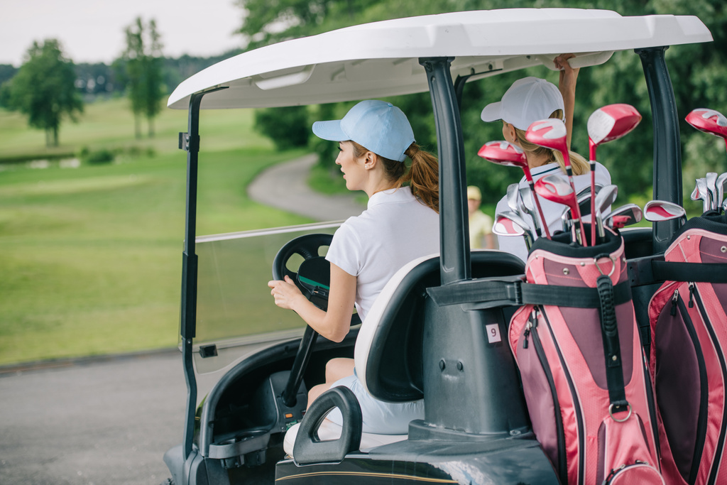 visão traseira de golfistas fêmeas em bonés montando carrinho de golfe com equipamento no campo de golfe
 - Foto, Imagem