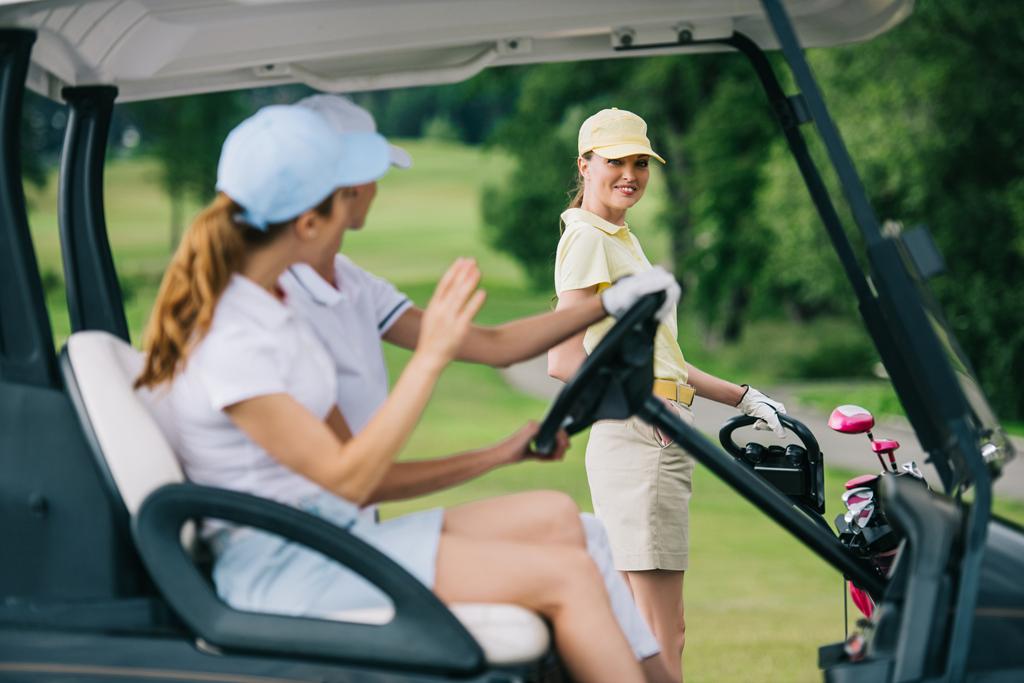 selective focus of female golfers in caps in golf cart greeting friend with golf gear at golf course - Photo, Image