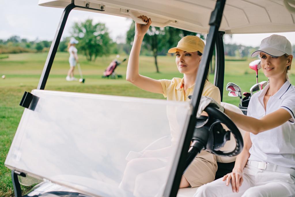 smiling female golfers in caps riding golf cart at golf course - Photo, Image