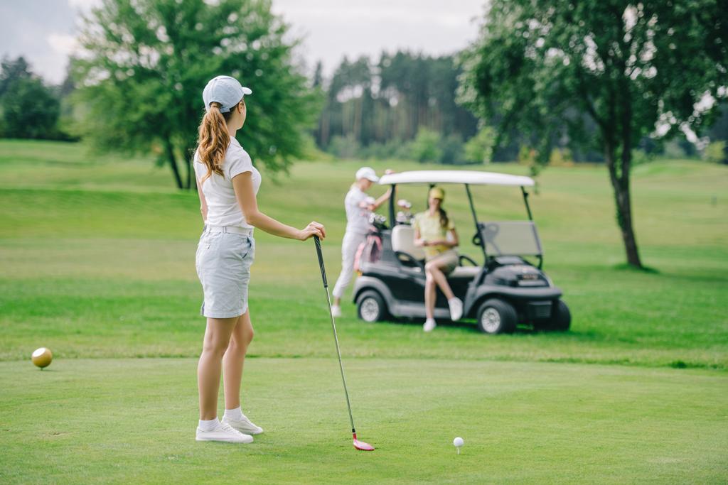 selectieve aandacht van vrouw met Golfclub kijken naar vrienden rustend op de golfkar op groen gazon - Foto, afbeelding