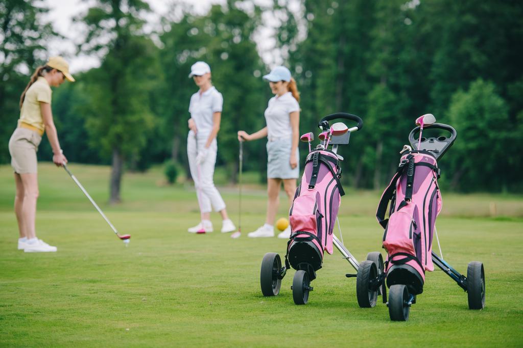 enfoque selectivo de equipo de golf, mujer en gorra jugando al golf y amigos de pie cerca en el campo de golf
 - Foto, Imagen