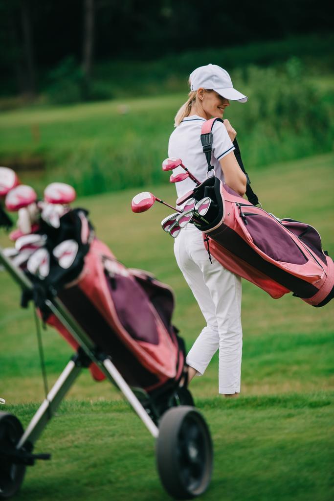 улыбающаяся женщина в поло и кепке с снаряжением для гольфа на поле для гольфа в летний день
 - Фото, изображение