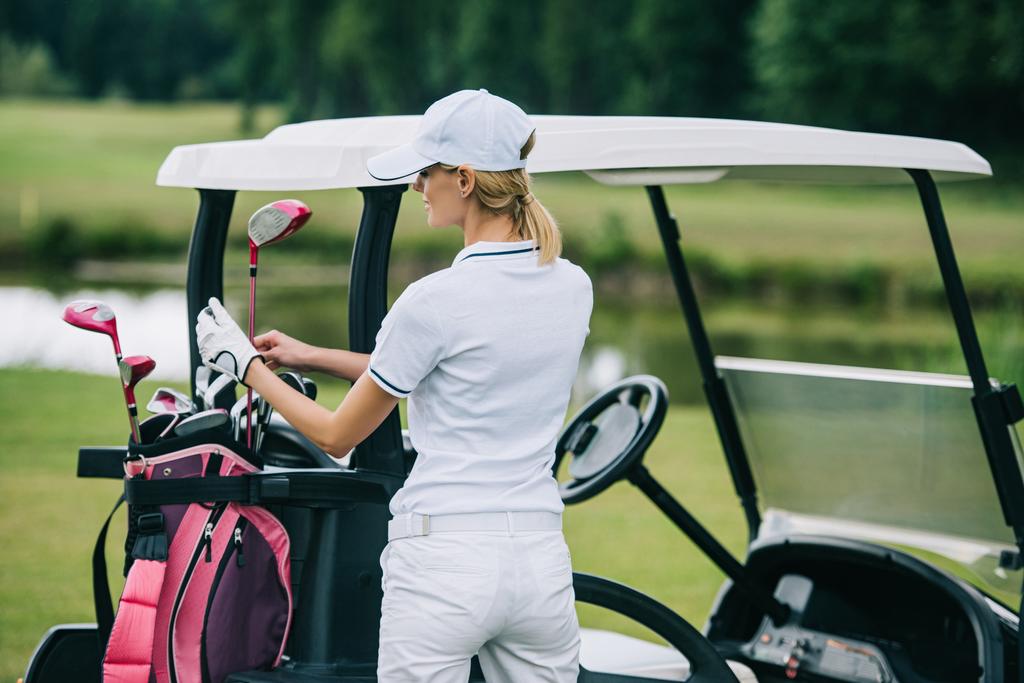 Achteraanzicht van de vrouw in polo en cap met golf gear staande op de golfkar op golfbaan op zomerdag - Foto, afbeelding