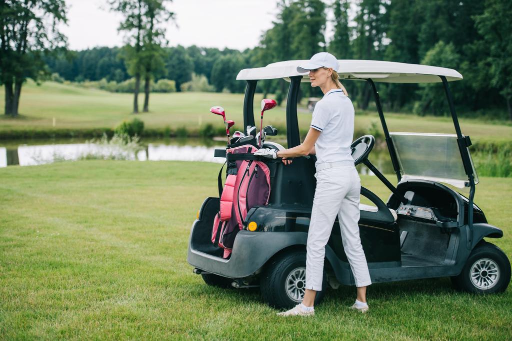 Frau in Polo und Mütze mit Golfausrüstung steht an einem Sommertag am Golfplatz am Golfcart - Foto, Bild