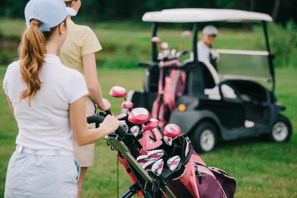 selective focus of female golf players with golf equipment and friend in golf cart behind on green lawn - Photo, Image