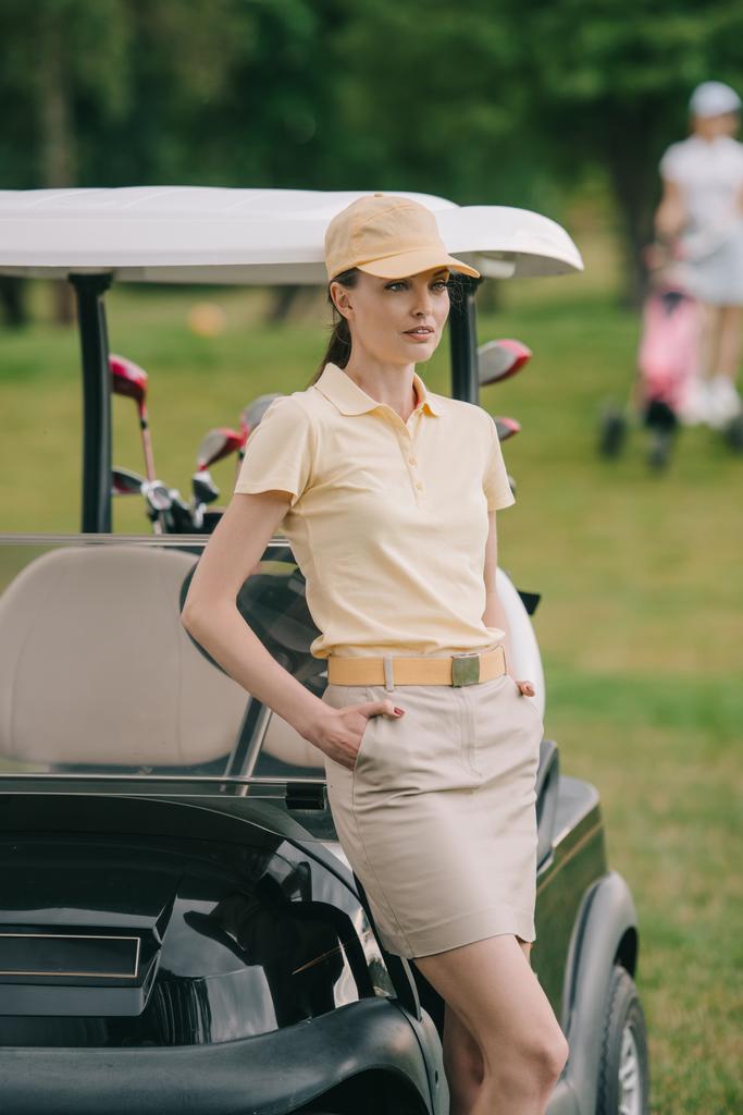 selective focus of female golf player in cap with hands in pockets at golf cart on green lawn - Photo, Image