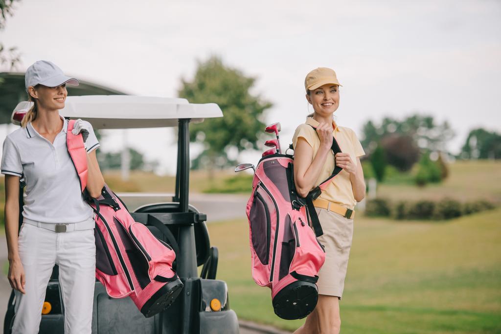 hymyilevät naiset pooloissa ja lippiksissä golflaitteilla golfkentällä kesäpäivänä
 - Valokuva, kuva