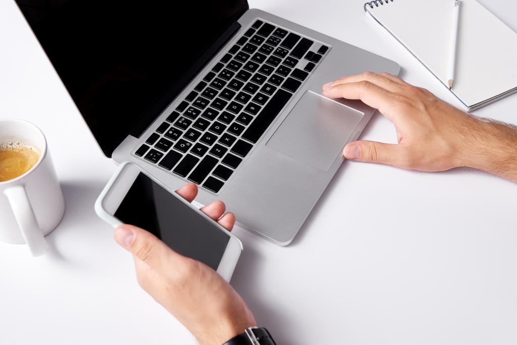 обрізаний знімок людини, що працює з ноутбуком і тримає смартфон з порожнім екраном на білій поверхні
 - Фото, зображення