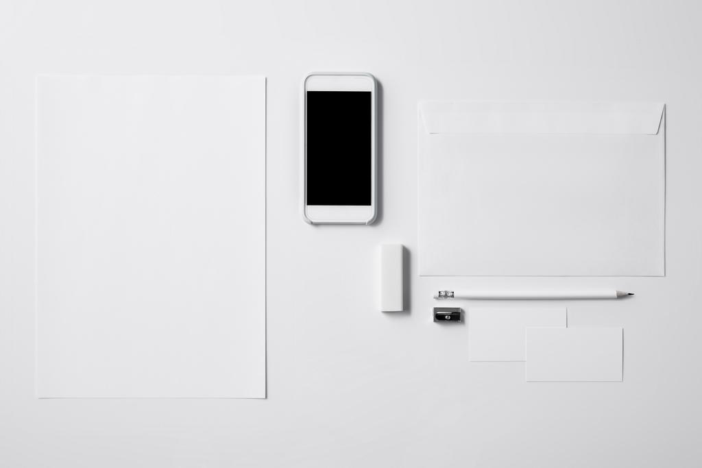 pohled shora na smartphone s prázdnou obrazovku a kancelářské potřeby na bílém stole pro makety - Fotografie, Obrázek