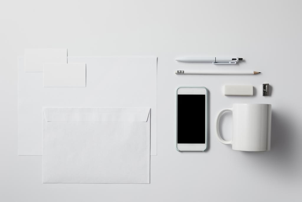 vista superior do smartphone com vários suprimentos e papéis em branco na superfície branca para mockup
 - Foto, Imagem