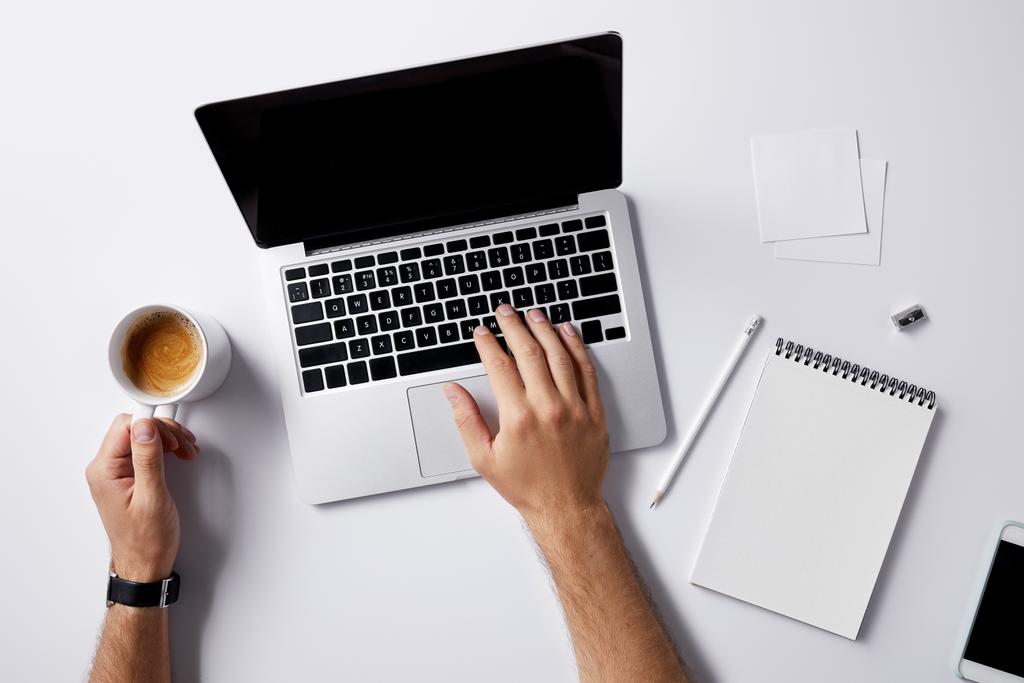 обрезанный снимок человека, работающего с ноутбуком и пьющего чашку кофе на рабочем месте на белой поверхности для макета
 - Фото, изображение