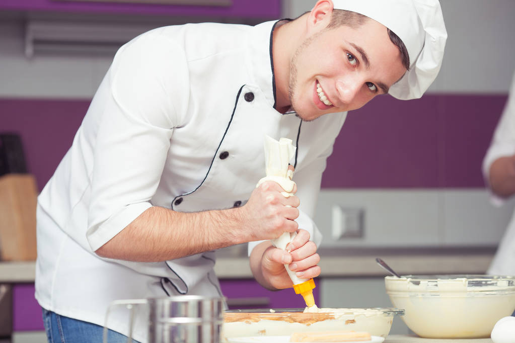 Tiramisu, gotowanie, przekazując koncepcja egzaminu. Portret uśmiechający się stażysta w mundurze kucharz Dokonywanie włoski deser w nowoczesnej kuchni. Wewnątrz strzał - Zdjęcie, obraz